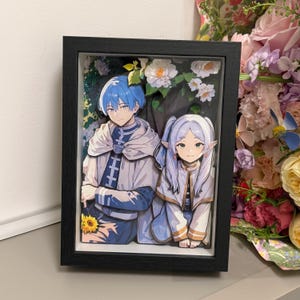Op de afbeelding: Ingelijste kunstwerk met twee anime-achtige figuren in blauwe en witte kleding. Het mannelijke personage, met blauw haar, houdt een zonnebloem vast. Het vrouwelijke personage heeft lang wit haar. Het kunstwerk is geplaatst tegen een achtergrond van witte bloemen en groen.