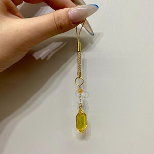 Könnte beinhalten: Ein goldfarbener Handy-Anhänger mit einem gelben Kristallanhänger. Der Anhänger hat einen geflochtenen Abschnitt, einen kleinen Ring und klare und orangefarbene Perlen. Der Kristall ist leuchtend gelb und setzt einen Farbakzent.