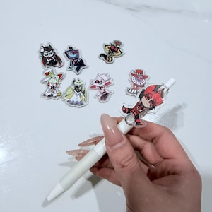 Puede incluir: Un bolígrafo blanco con un dije de personaje de dibujos animados adjunto, rodeado de otros dijes de personajes. Los dijes presentan varios diseños coloridos y probablemente son de acrílico o plástico. El bolígrafo lo sostiene una persona.