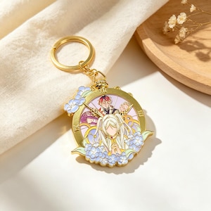May include: A gold-toned metal keychain featuring a circular clock-face design with Roman numerals. The center displays an anime-style illustration of two characters with white and red hair, surrounded by light blue floral accents and gold detailing.