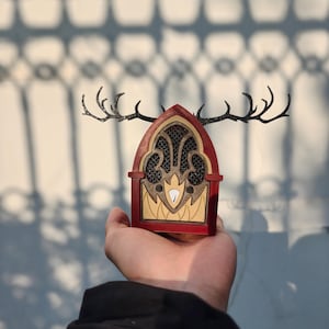 Könnte beinhalten: Ein dekoratives Holzradio mit rotem Rahmen, aufwendigen beige-braunen Details und einem schwarzen Geweihdesign. Das Radio wird in einer Hand gehalten und zeigt sein einzigartiges, künstlerisches Design.