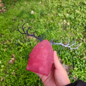 May include: A red, rectangular object with black antlers attached. The antlers are made of a thin, dark material and are curved. The object is held in a hand against a background of green grass.
