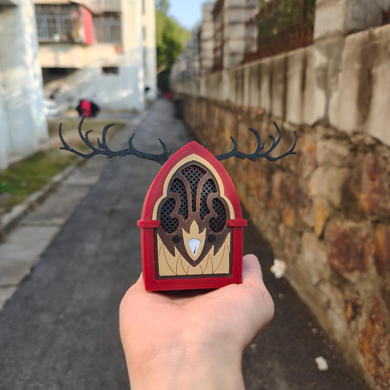 May include: A red and brown decorative radio with a deer antler design on top. The radio has a speaker grill and a gold-colored emblem at the bottom. The radio is held in a hand.