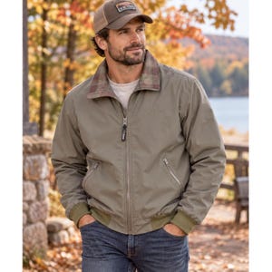 May include: A sage green zip-up jacket with a plaid-lined collar, ribbed cuffs, and two zippered side pockets. The jacket features a full-length front zipper with a branded pull tab and is worn over a white shirt with blue denim jeans.