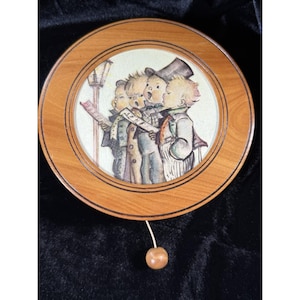 May include: A circular wooden music box with a painted image of four children singing carols. The image is framed by a light brown wooden border. A small wooden ball hangs from the bottom. The music box is a vintage item.