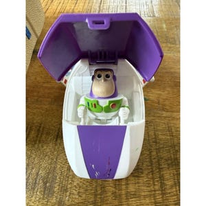 Puede incluir: Un juguete de nave espacial de plástico blanco y morado con la escotilla de la cabina abierta. Dentro de la cabina hay una pequeña figura de acción de Buzz Lightyear con un traje blanco, verde y morado. El exterior tiene un panel frontal morado.