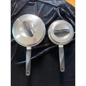 May include: Two stainless steel saucepans with lids. The pans have black handles and lids with black handles. The pans are of different sizes, with the larger one on the left and the smaller one on the right. The pans are on a dark background.