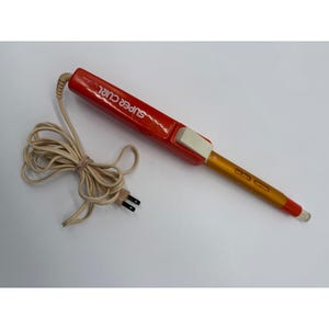 Puede incluir: Una plancha rizadora roja y dorada vintage con el texto "SUPER CURL by Calorie". La plancha rizadora tiene un interruptor blanco y un cable beige con un enchufe de dos clavijas. El cable está asegurado con una banda elástica marrón.