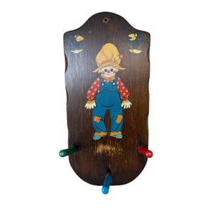Könnte beinhalten: Eine dunkelbraune Wandgarderobe aus Holz mit einer aufgemalten Vogelscheuche in der Mitte, flankiert von zwei schwarzen Krähen. Die Garderobe hat drei Haken in Grün, Rot und Blau sowie einen gewellten Rand mit einem Loch zur Aufhängung.