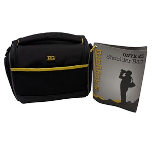 May include: A black shoulder bag with yellow trim and a top handle. The bag features a small black and yellow RG logo on the front. Beside the bag is a product tag labeled Ruggard Onyx 25 Shoulder Bag, featuring a silhouette of a person.