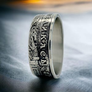 Op de afbeelding: Een zilveren ring met gegraveerde details en tekst. De ring heeft een gedetailleerd ontwerp met een historisch of artistiek thema. De tekst is in een vreemde taal. De ring wordt getoond op een getextureerd oppervlak.