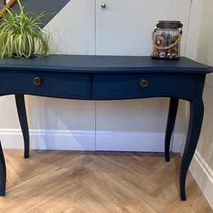 May include: A dark navy blue console table with two drawers and brass knobs. The table has curved legs and a curved front. A plant in a pot and a decorative jar sit on top of the table.