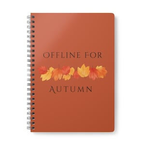 Pode incluir: Um caderno de espiral com uma capa de cor laranja queimado apresentando o texto preto "OFFLINE FOR AUTUMN" e uma linha horizontal de folhas de outono ilustradas em tons de amarelo, laranja e vermelho centrada entre as linhas de texto.