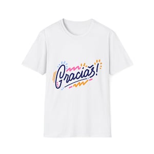Pode incluir: T-shirt branca com a palavra "Gracias!" em uma fonte colorida e estilizada. A palavra está em um estilo cursivo com detalhes em azul, laranja e rosa. O design está centralizado na frente da camiseta.
