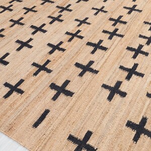 Könnte beinhalten: Ein beigefarbener Jute-Teppich mit einem sich wiederholenden Muster aus schwarzen Kreuzen und kurzen horizontalen Linien. Der Teppich hat eine gewebte Textur und eine neutrale Farbpalette, passend für verschiedene Einrichtungsstile.