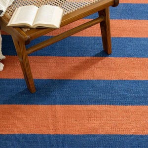 May include: A blue and orange striped rug with a woven texture. A wooden chair with a woven seat and a book are on the rug. A gray and white fringed throw is draped over the chair.