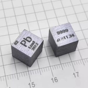 Puede incluir: Dos cubos metálicos de plomo. El primer cubo está grabado con 82, Pb, Lead y 207.2. El segundo cubo está grabado con 9999 y ρ=11.34. Los cubos están colocados sobre una superficie cuadriculada con una regla visible que muestra los números 15, 16 y 17.