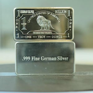 Op de afbeelding: Twee rechthoekige Duitse zilveren baren. De bovenste staaf toont een leeuw, de tekst "UNITED STATES OF AMERICA" en "ONE TROY OUNCE". De onderste staaf luidt ".999 Fine German Silver".