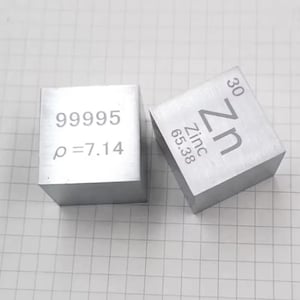 May include: Two silver-colored metal cubes. One cube features the text 99995 and ρ=7.14. The other cube displays the chemical symbol Zn, the word Zinc, the atomic number 30, and the atomic mass 65.38 engraved on the top surface of the metal block.