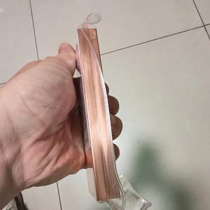 May include: A rectangular object with a metallic rose gold finish encased in a clear plastic blister pack. The item features a smooth, reflective surface and a slim profile, with a small clear plastic loop at the top for hanging display purposes.