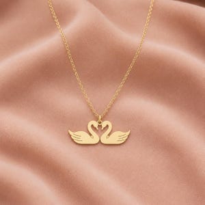 May include: A gold-toned necklace featuring a pendant shaped like two swans facing each other, with their necks curved to form a heart shape. The pendant is suspended from a delicate, thin gold-toned chain with a simple link design.