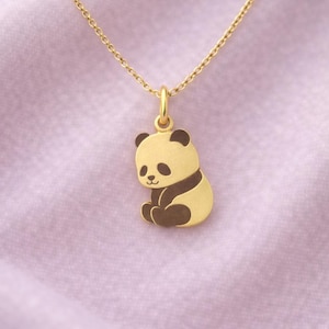 Op de afbeelding: Een gouden panda beer hanger ketting. De panda heeft zwarte accenten op zijn oren, ogen en lichaam. De ketting is ook goudkleurig en hangt tegen een zachte, roze achtergrond.