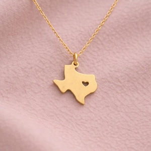 May include: A gold-coloured necklace featuring a pendant in the shape of Texas, with a heart-shaped cutout. The delicate gold chain hangs against a pink backdrop. The pendant is approximately 2.5 cm in length.