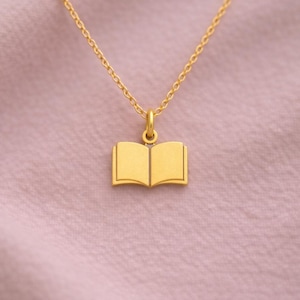 May include: A gold-colored necklace featuring a pendant shaped like an open book. The pendant is a detailed miniature of an open book. The necklace chain is also gold and hangs against a pink background.