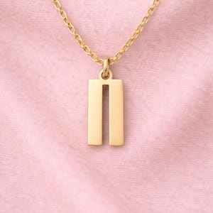 Puede incluir: Un collar dorado con un colgante rectangular con dos barras verticales. El colgante cuelga de una delicada cadena. El fondo es una tela rosa suave.