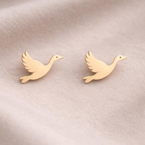 Puede incluir: Un par de pendientes de botón dorados con forma de pájaro. Los pendientes tienen la forma de un pájaro en vuelo, con las alas extendidas y un pequeño pico. Los pendientes se muestran sobre una tela beige claro.