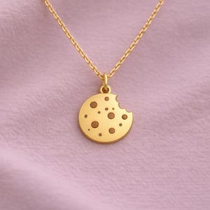 Puede incluir: Collar dorado con colgante de galleta. El colgante redondo tiene un mordisco y manchas marrones. La cadena también es dorada, sobre un fondo de tela rosa claro. Una joya encantadora.
