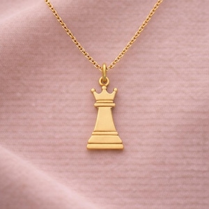 Peut inclure: Un collier pendentif doré représentant une pièce d'échecs, la reine. Le pendentif est une représentation stylisée et détaillée d'une reine, suspendue à une chaîne délicate. Le fond est un tissu rose doux.