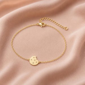 May include: A gold-toned metal chain bracelet featuring a small, circular charm shaped like a cookie with a bite taken out of the side. The charm includes several small, circular indentations on its surface. The bracelet has a lobster claw clasp.