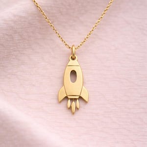 Puede incluir: Un collar con colgante de cohete dorado. El colgante presenta un diseño detallado de cohete con una ventana ovalada y tres llamas de escape. La cadena del collar es delicada y dorada, sobre un fondo rosa suave.
