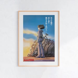 Puede incluir: Un póster enmarcado con un personaje de una película de animación, de perfil contra un cielo de puesta de sol. El personaje viste un atuendo morado claro y sostiene un arma. Texto japonés visible en la parte superior e inferior del póster.