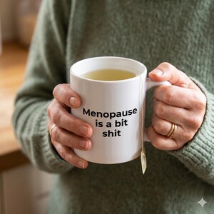 May include: White ceramic mug filled with tea, held by a person wearing a green sweater. The mug has the text "Menopause is a bit shit" printed in black. A tea bag is visible.