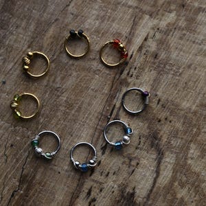 Può includere: Una collezione di piercing per il naso in tonalità oro e argento, ognuno ornato da perline di vari colori, tra cui nero, rosso, verde e blu. Gli anelli sono disposti su una superficie di legno rustico.
