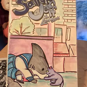 May include: Comic book cover titled "Sailor Jeff and the Land Shark." The artwork features a shark in a sailor outfit interacting with a gray cat. The background includes a building facade and the comic book is displayed in a clear stand.