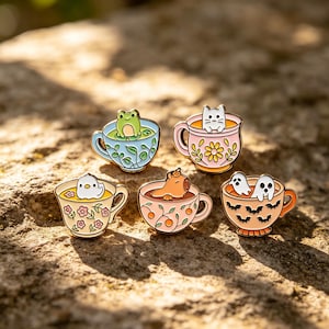 May include: A collection of enamel pins featuring whimsical creatures in teacups. The pins include a frog, cat, bird, capybara, and ghosts. Each teacup has a unique color and design, with floral and bat motifs.