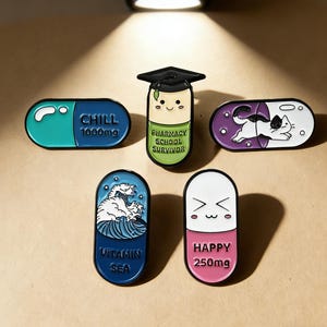 May include: A collection of five enamel pins in the shape of pills. The pins feature various designs, including a cat, a wave, and text that reads "CHILL 1000mg", "PHARMACY SCHOOL SURVIVOR", "VITAMIN SEA", and "HAPPY 250mg".
