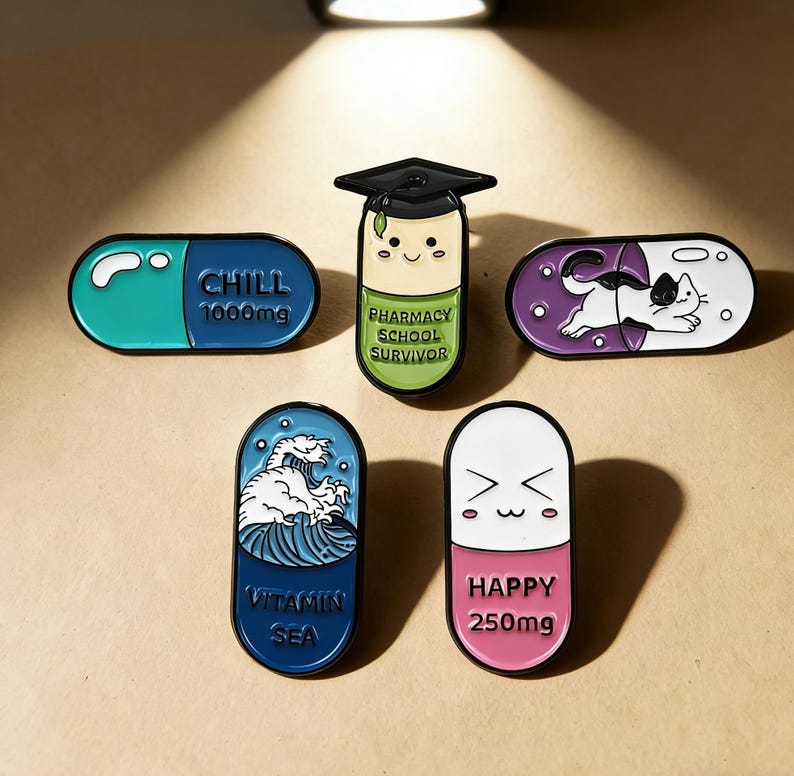 May include: A collection of five enamel pins in the shape of pills. The pins feature various designs, including a cat, a wave, and text that reads "CHILL 1000mg", "PHARMACY SCHOOL SURVIVOR", "VITAMIN SEA", and "HAPPY 250mg".