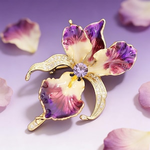 May include: A gold-toned brooch in the shape of an iris flower, featuring purple and cream-colored petals. The brooch is adorned with small, sparkling crystals and a central purple gemstone. The design is detailed and elegant, with a pin closure on the back.