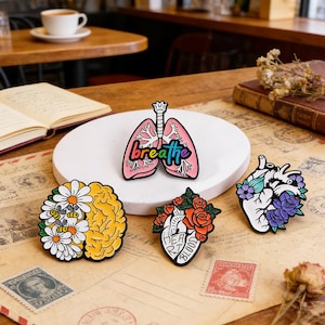 Könnte beinhalten: Eine Sammlung farbenfroher Emaille-Anstecknadeln mit anatomischen Designs. Enthält ein Paar rosa Lungen mit dem Wort "breathe", ein Gehirn mit Gänseblümchen, ein Herz mit Rosen und ein Herz mit lila Blumen. Auf einem Hintergrund im Vintage-Stil.
