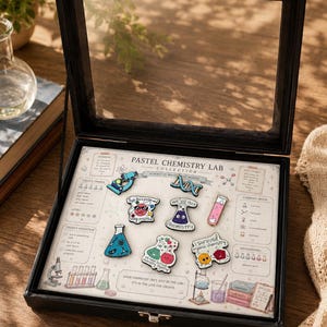 Puede incluir: Una vitrina negra con tapa de cristal, que muestra una colección de pines de temática química en colores pastel. Los pines presentan varios equipos de laboratorio y frases como "Stay positive" y "We've got chemistry". La vitrina está abierta, revelando un fondo impreso.