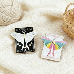 May include: Two enamel pins featuring butterfly designs. One pin has a black background with a white butterfly and the words "yes" and "no". The other pin has a white background with a colorful butterfly and the words "yes" and "no".
