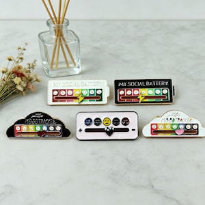 May include: A collection of enamel pins with mood tracker and social battery designs. The pins feature a range of colors, including red, yellow, green, and black, with text that reads "MY SOCIAL BATTERY" and "MOODTRACKER".