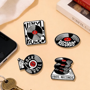 以下が含まれることがあります： ビニールレコードのデザインの4つのエナメルピン。1つは「Vinyl Rules」と書かれた長方形です。もう1つは「Records」と書かれた楕円形です。3つ目は「Rock & Roll」と書かれたレコードプレーヤーです。最後は「Vinyl Records」と書かれた積み重ねられたレコードです。