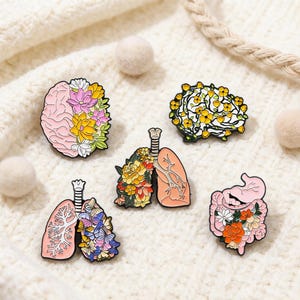 Könnte beinhalten: Fünf Emaille-Anstecknadeln mit anatomischen Designs und floralen Akzenten. Die Anstecknadeln zeigen ein Gehirn, Lungen und einen Magen, jeweils mit bunten Blumen verziert. Die Anstecknadeln sind vor einem cremefarbenen, strukturierten Hintergrund platziert.