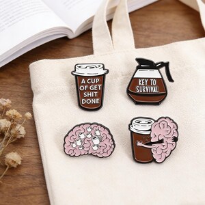 Könnte beinhalten: Vier Emaille-Anstecknadeln mit Kaffeemotiven. Eine Anstecknadel ist eine Kaffeetasse mit dem Text "A CUP OF GET SHIT DONE". Eine andere ist eine Kaffeekanne mit dem Text "KEY TO SURVIVAL". Die anderen beiden Anstecknadeln zeigen ein Gehirn und Kaffeetassen.
