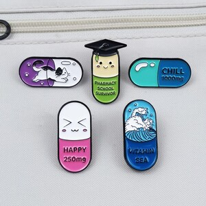 May include: A collection of enamel pins in the shape of pills. Designs include a cat, a graduate, and pills with the words "CHILL", "HAPPY", and "VITAMIN SEA". The pins are in various colors, including purple, blue, and pink.